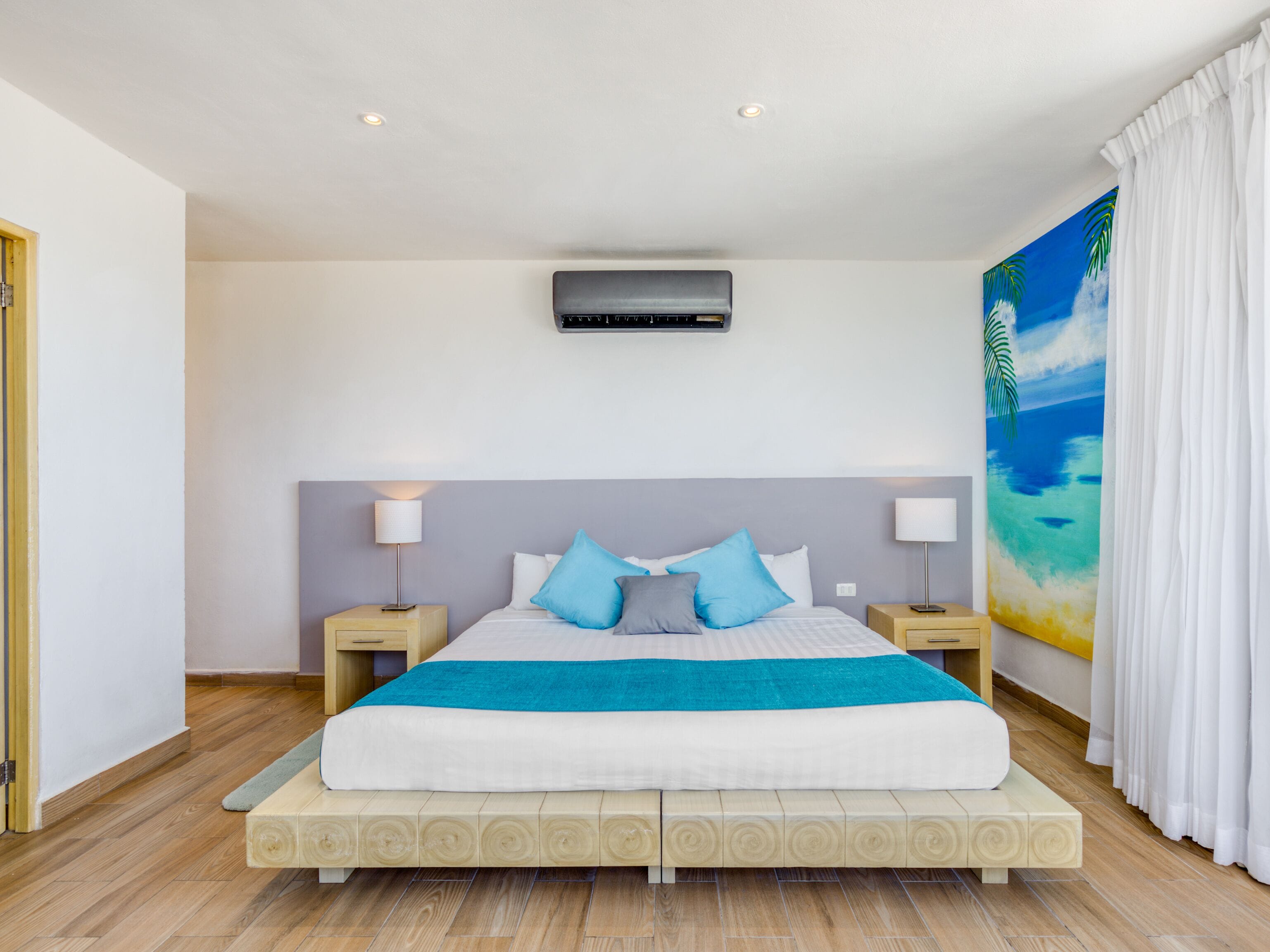 A modern bedroom with a large bed, two bedside tables with lamps, a ceiling air conditioner, and a tropical-themed painting on the wall.