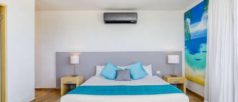 A modern bedroom with a large bed, two bedside tables with lamps, a ceiling air conditioner, and a tropical-themed painting on the wall.