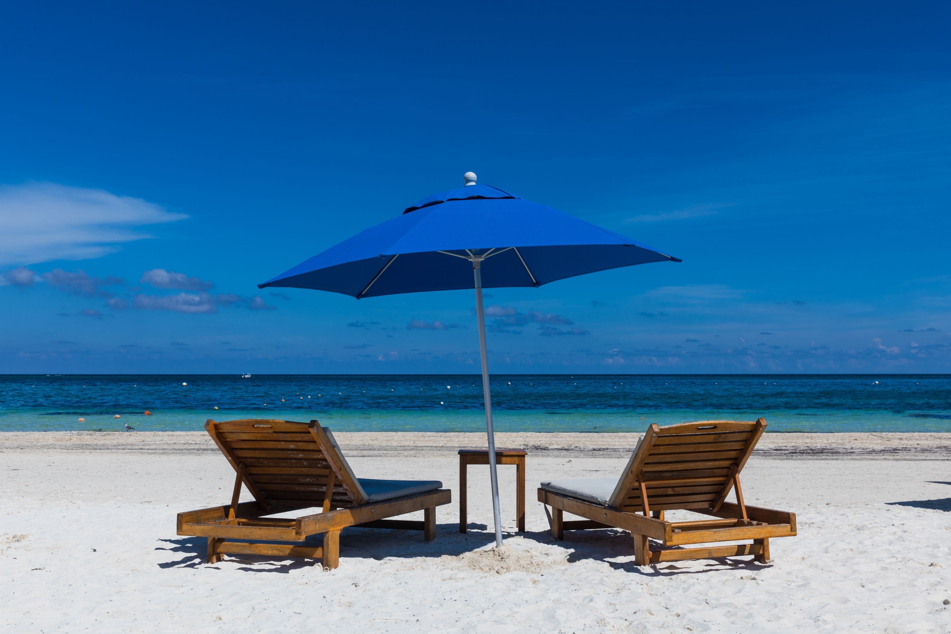 Dos sillones de madera y una sombrilla azul sobre una playa arenosa, con un cielo azul despejado y el océano al fondo.