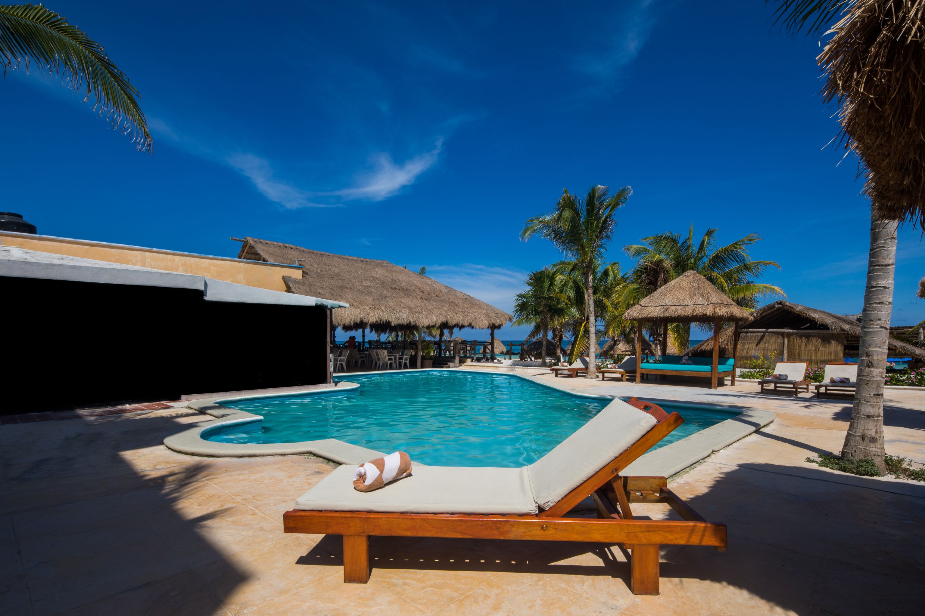 Un área de piscina con sillas de descanso, palmeras y estructuras de techo de paja.