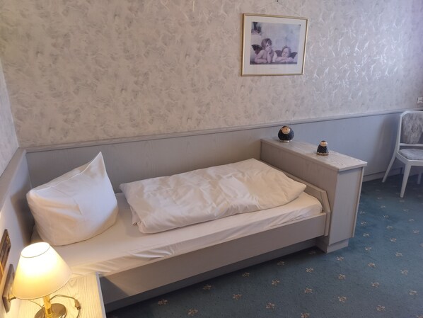 Una cama pequeña y bien hecha con sábanas blancas en una habitación con pared texturizada y un cuadro enmarcado encima.