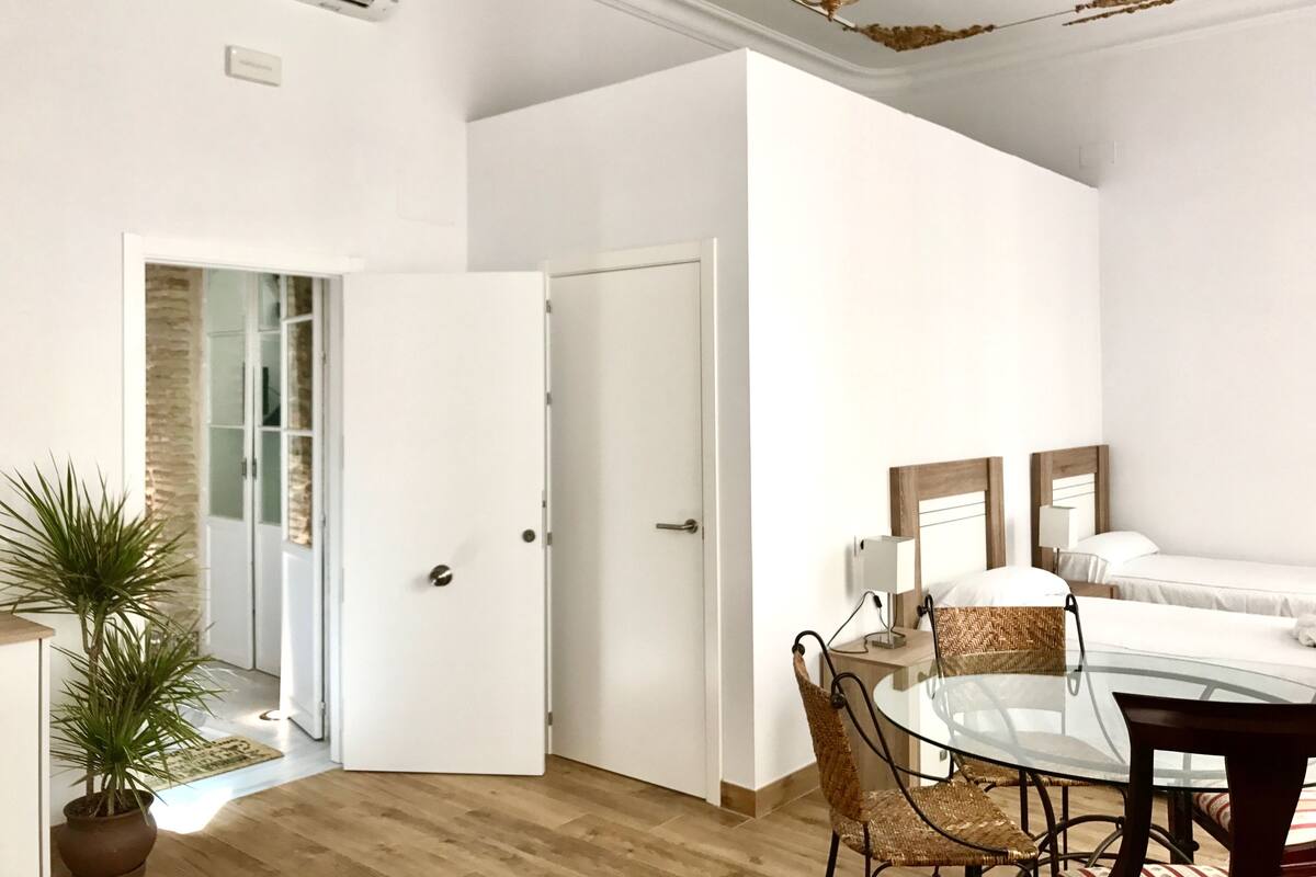 A modern bedroom with a glass table, wicker chairs, a bed with white linens, and a door leading to a bathroom.