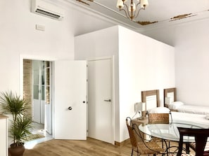 A modern bedroom with a glass table, wicker chairs, a bed with white linens, and a door leading to a bathroom.