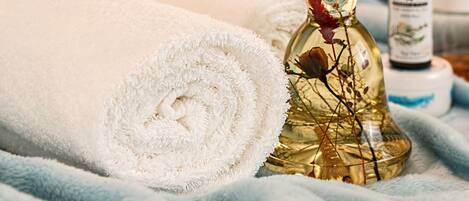 A rolled white towel, a glass bottle with dried flowers, and a small bottle with a label, all on a blue textured surface.