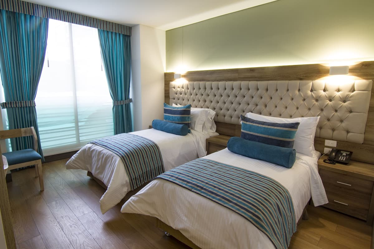 Quarto de hotel com duas camas, cabeceira de madeira, mesa de cabeceira com telefone e uma cadeira.
