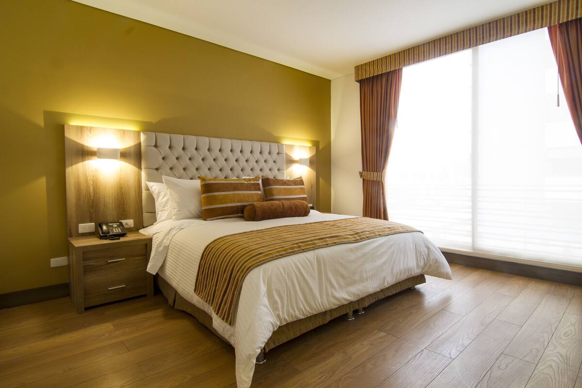Quarto de hotel com uma cama grande, mesa de cabeceira de madeira com telefone e janela com cortinas.