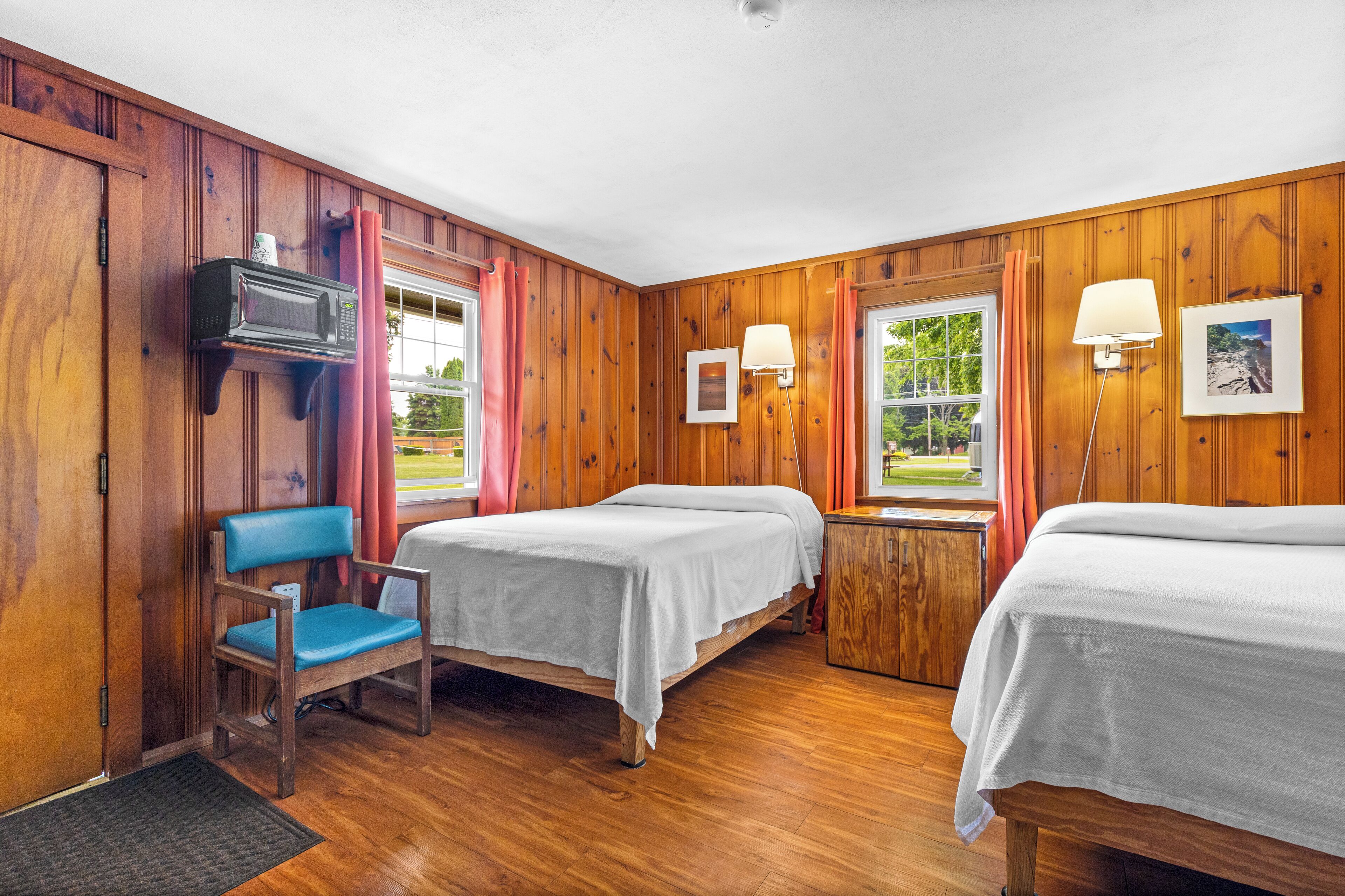 A room with wooden paneling, two beds with white linens, a chair, a TV, and a lamp.