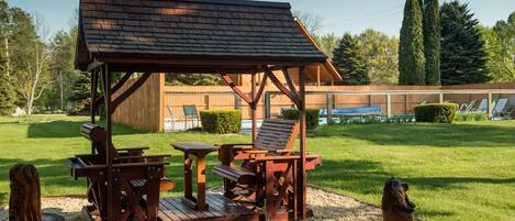 A wooden outdoor seating area with a table and chairs, surrounded by a gravel path and greenery.