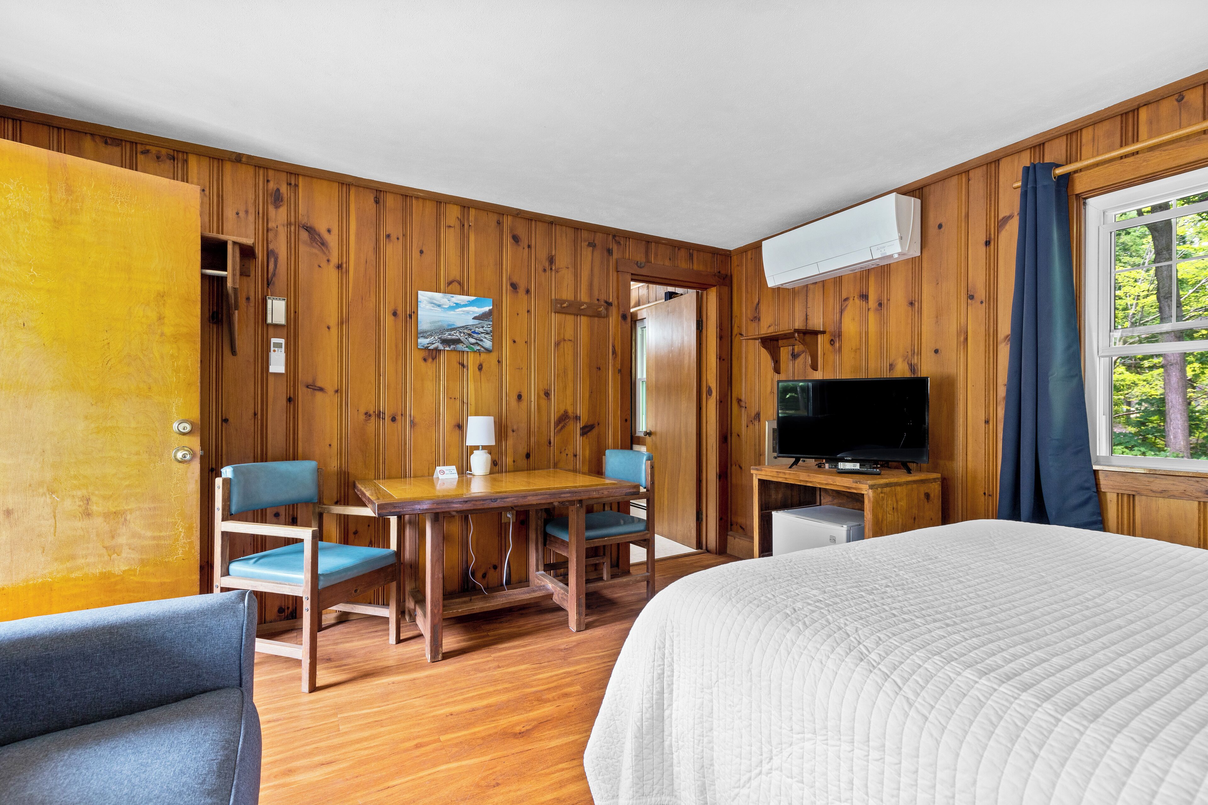 A wooden-paneled room with a bed, a television, a desk with chairs, and a window with curtains.