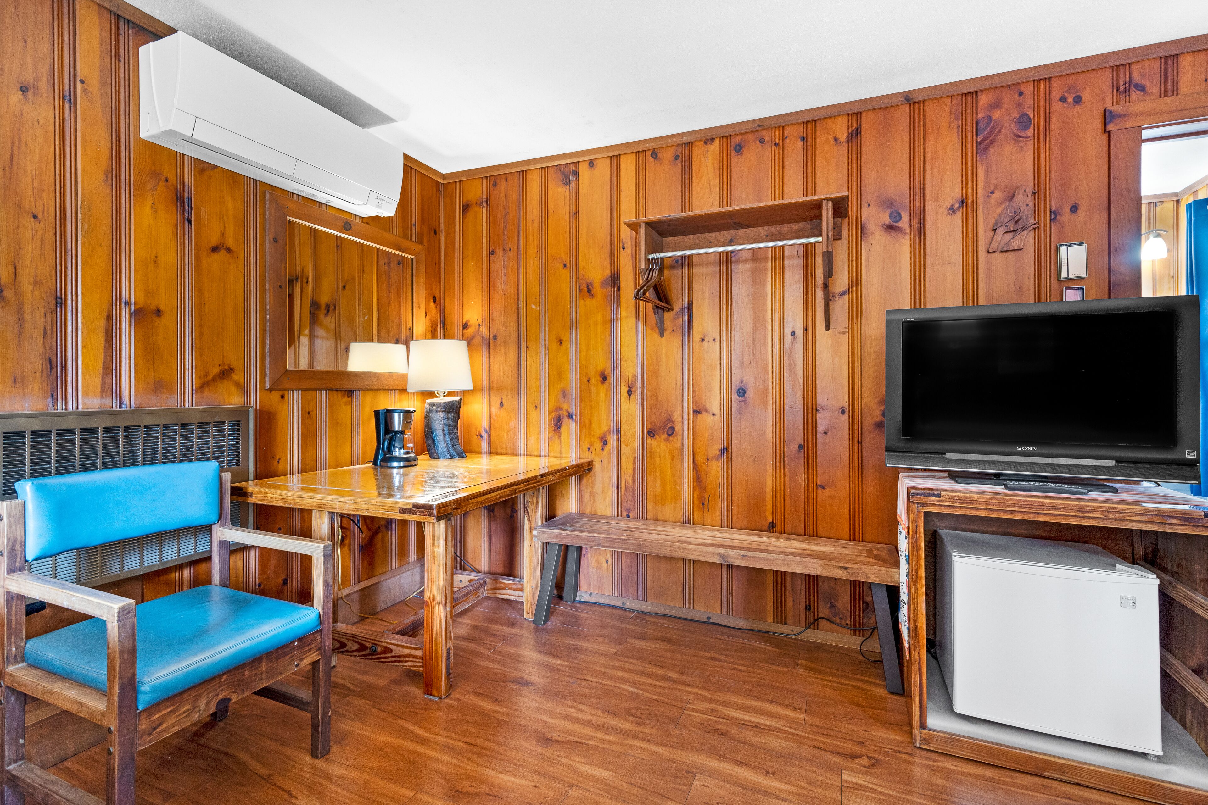 A wooden-paneled room with a wooden table, a blue chair, a bench, a television, and a small white refrigerator.