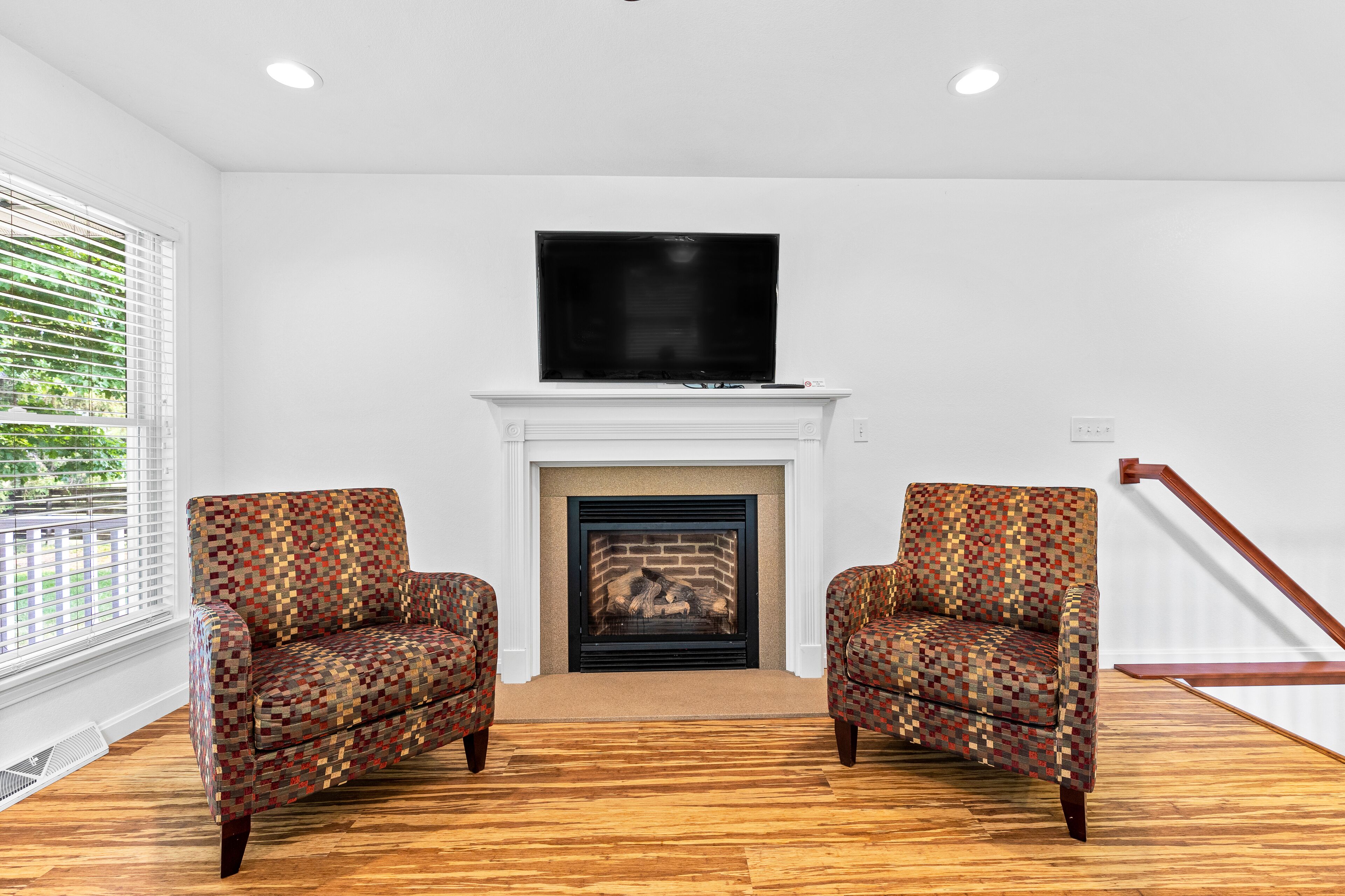 A living room with a fireplace, two patterned armchairs, a flat-screen TV above the fireplace, and a wooden floor.