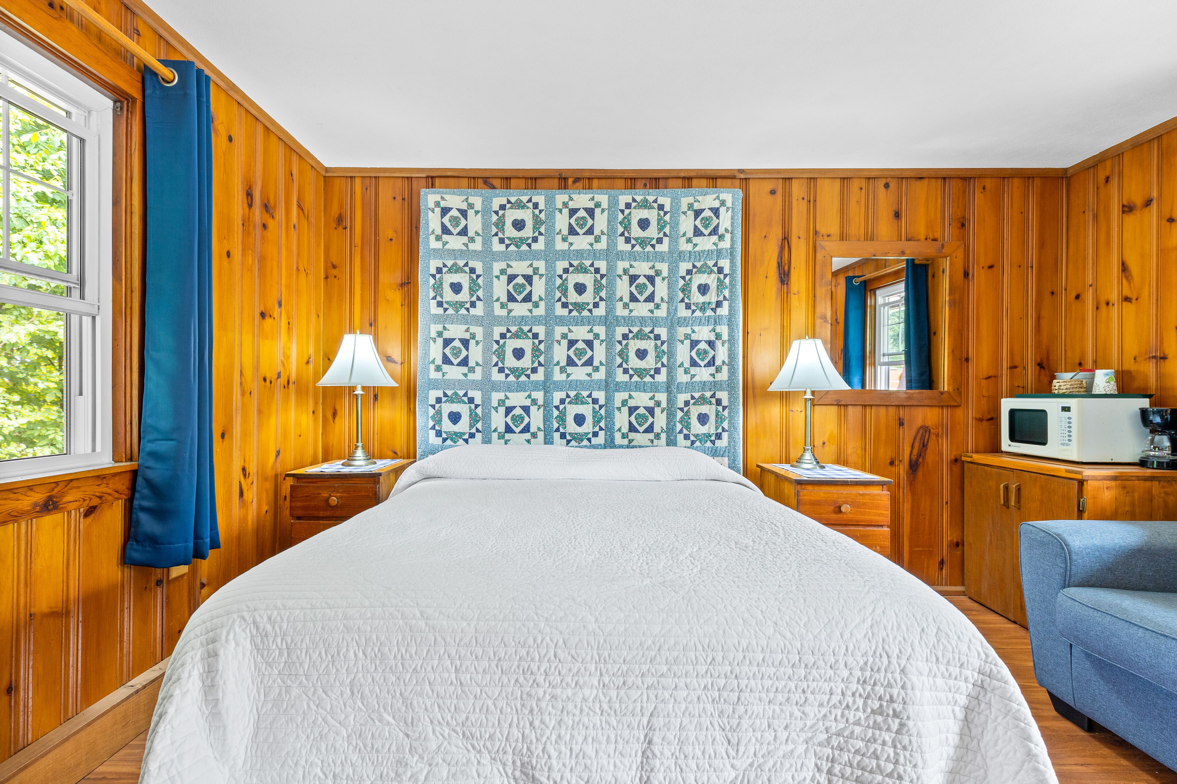 A room with wooden walls, a bed with a white quilt, two bedside tables with lamps, a blue chair, a microwave, and a window with blue curtains.
