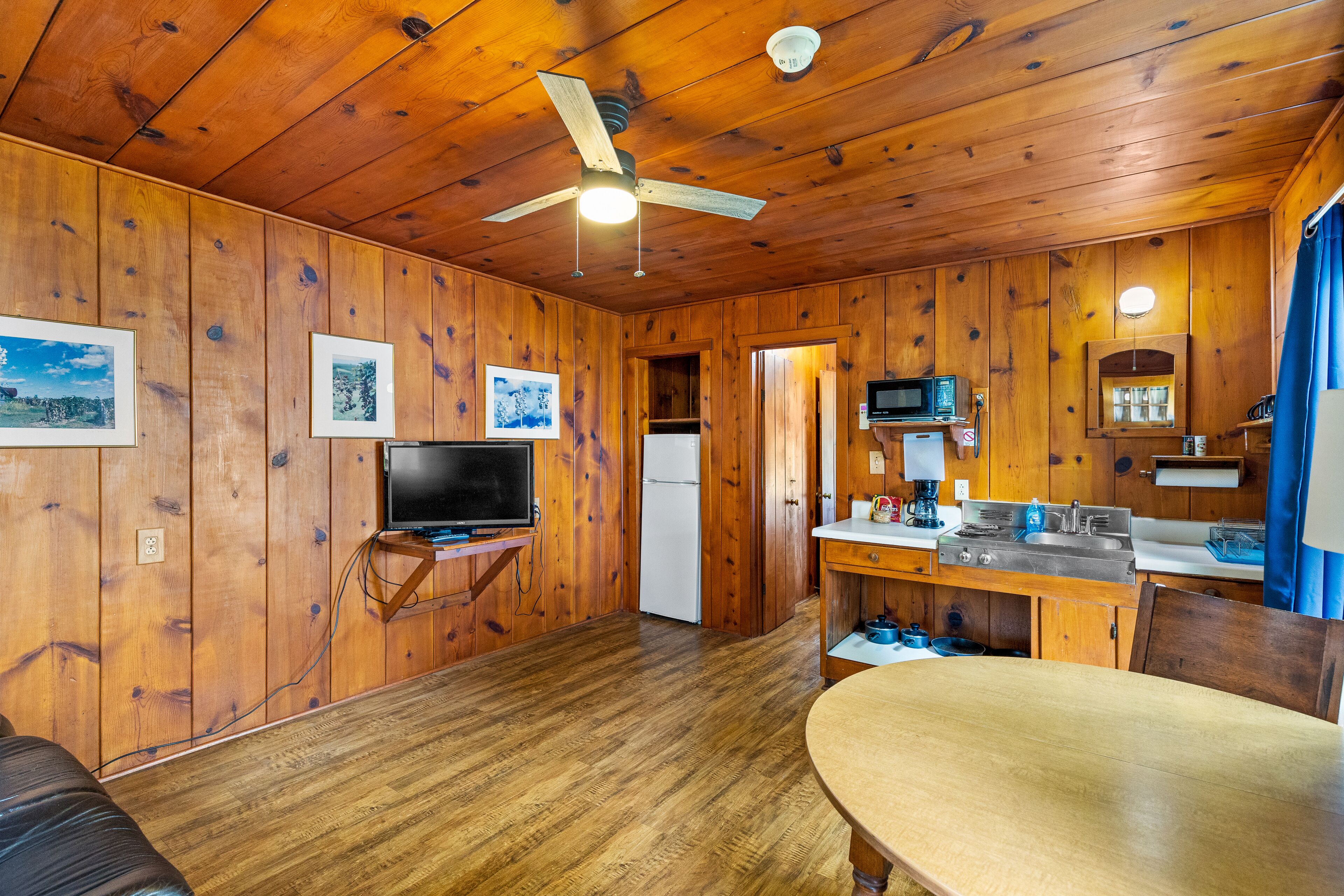 A wooden cabin interior with a dining table, kitchen area, ceiling fan, and mounted television.