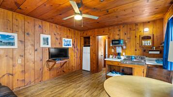 A wooden cabin interior with a dining table, kitchen area, ceiling fan, and mounted television.