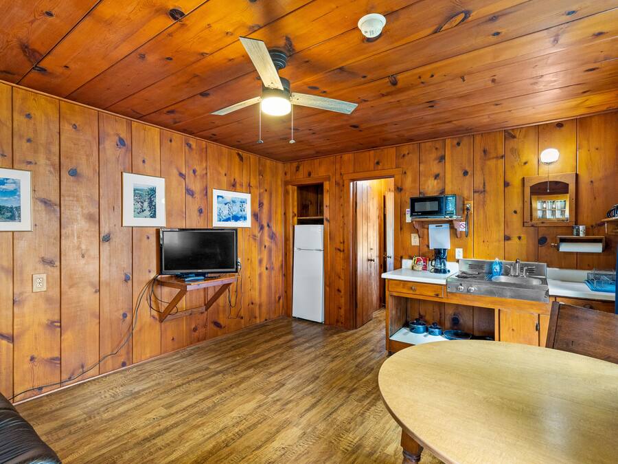 A wooden cabin interior with a dining table, kitchen area, ceiling fan, and mounted television.
