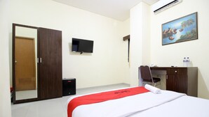 A hotel room with a bed, a desk, a chair, a wardrobe, a television, and a painting on the wall. - RedDoorz @ Swakarya Kaliurang (Depok)