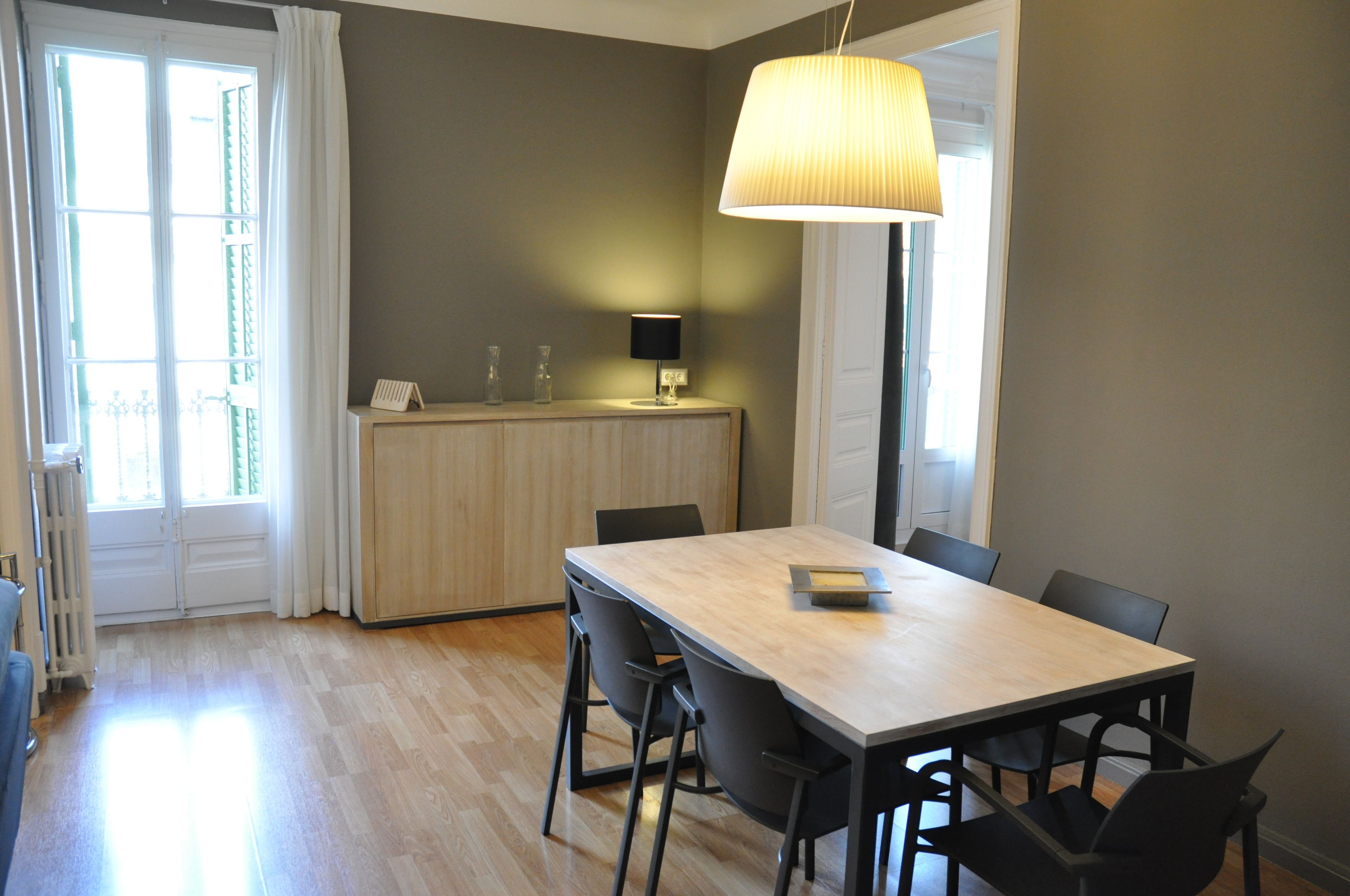 Un comedor con una mesa de madera y sillas negras, una lámpara colgante y vistas a un balcón a través de una ventana con cortinas blancas.