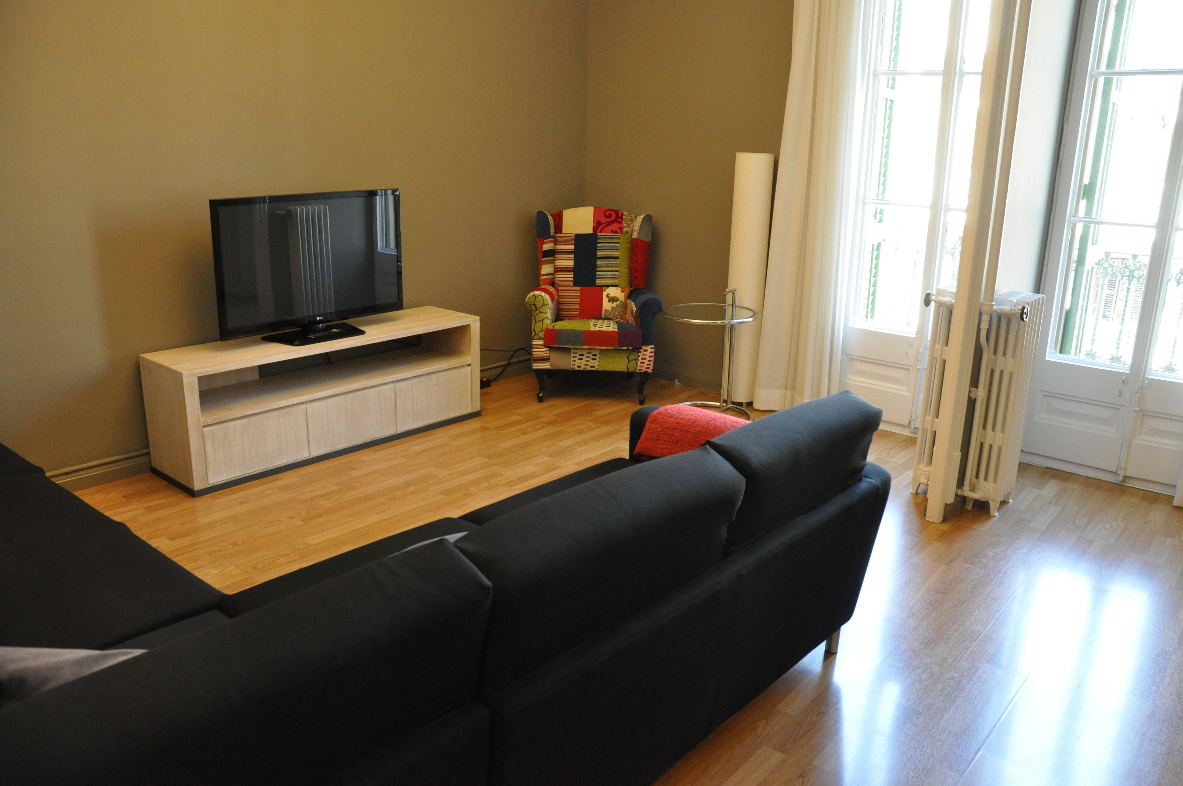 A living room with a flat-screen TV on a wooden stand, a colorful armchair, a black sofa, and a radiator near a window.