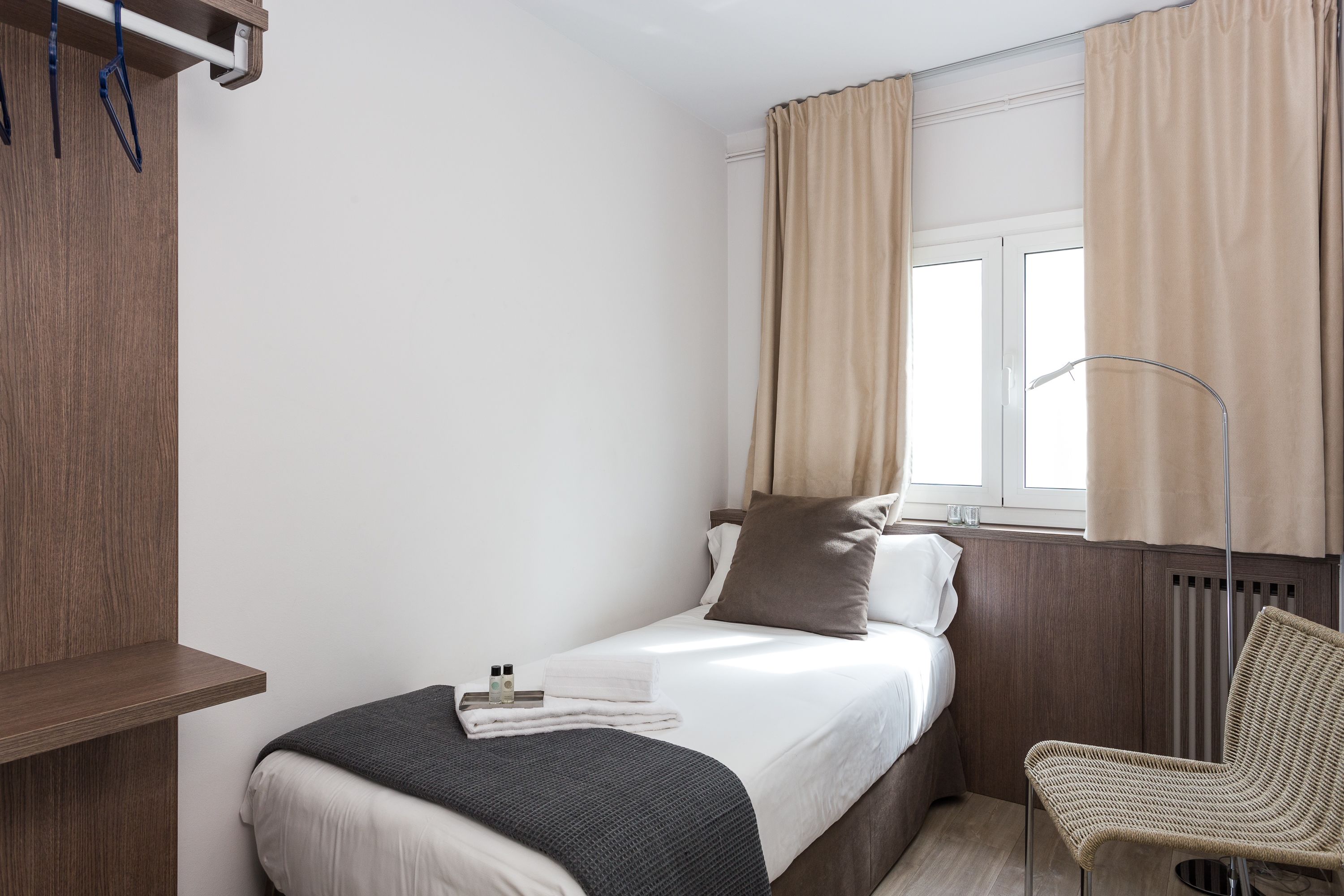 A small, neatly arranged bedroom with a bed, a chair, a shelf, and a window with curtains.