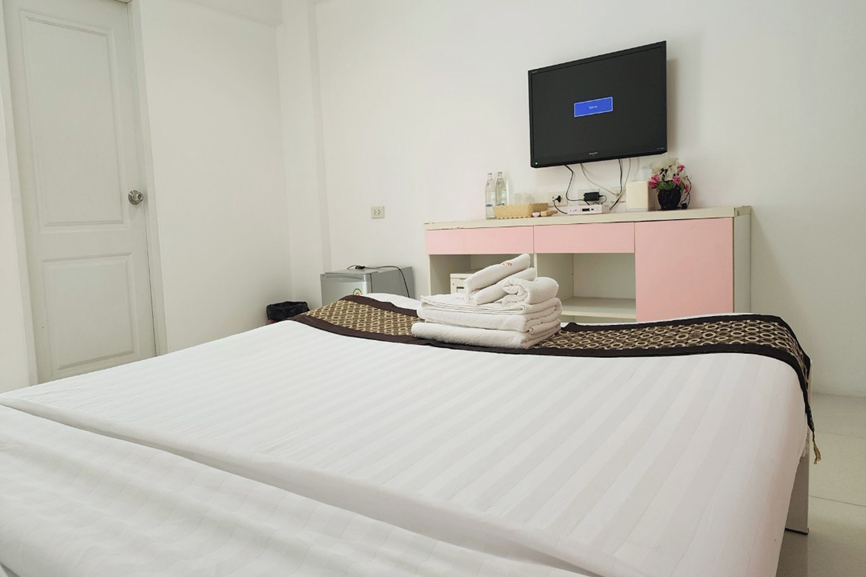 A hotel room with a bed, a TV mounted on the wall, a desk with a computer, and a pink and white minibar.