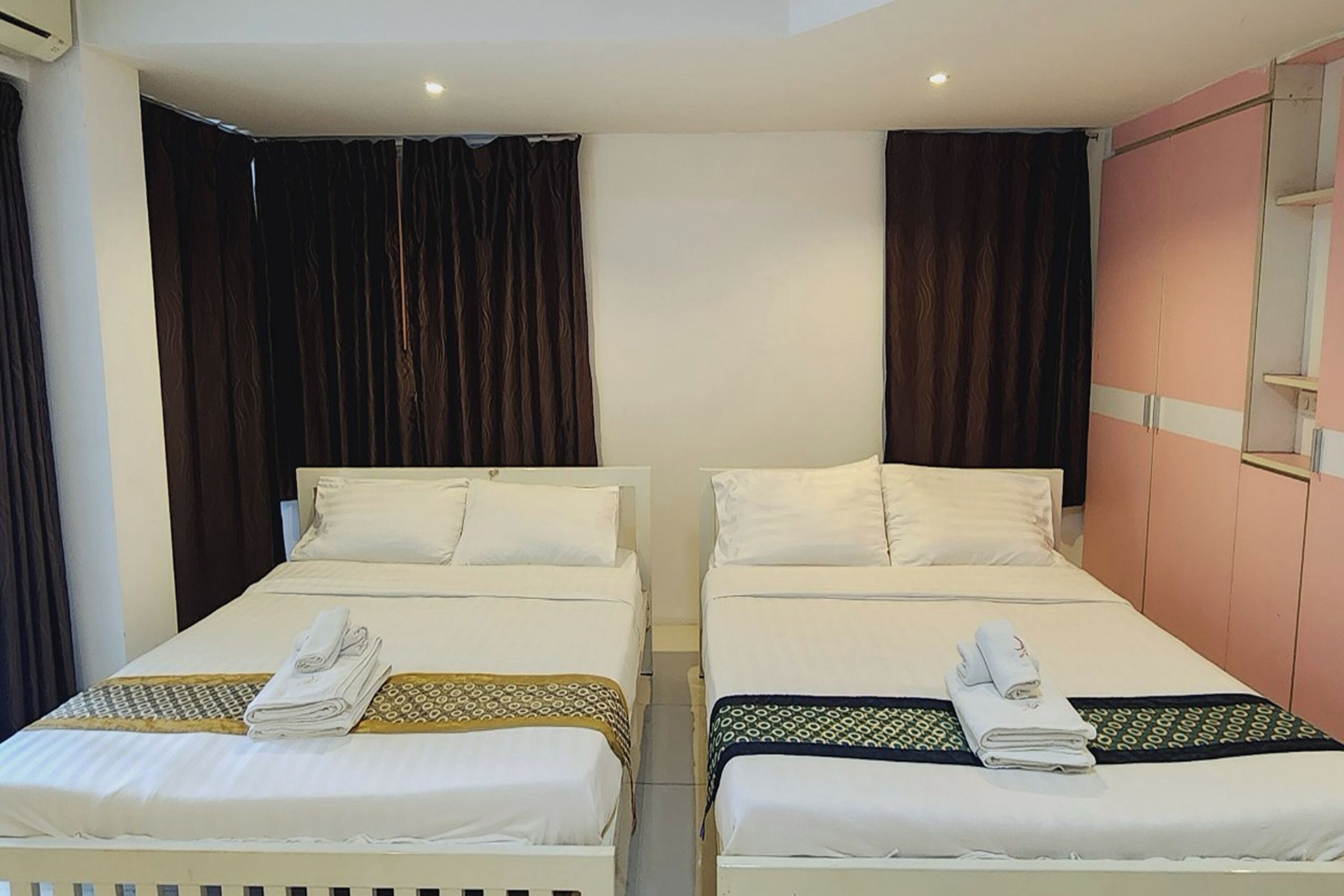 A hotel room with two single beds, each with a white mattress and pillow, and a patterned bedspread. There is a wardrobe with a mirror and a pink panel on the right side.