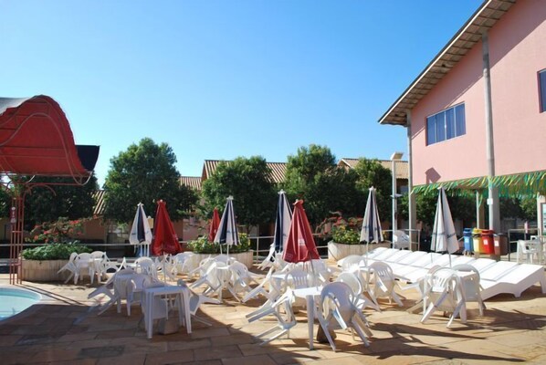 A poolside area with white lounge chairs and umbrellas, a swimming pool, and a building with a pink facade. - Apartment diRoma Fiori - Achei Férias (Caldas Novas)