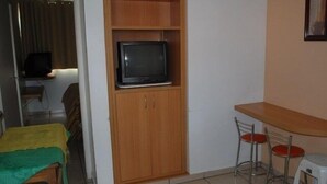 A small, compact living space with a wooden cabinet containing a television, a dining table with two chairs, and a small stool. - Apartment diRoma Fiori - Achei Férias (Caldas Novas)