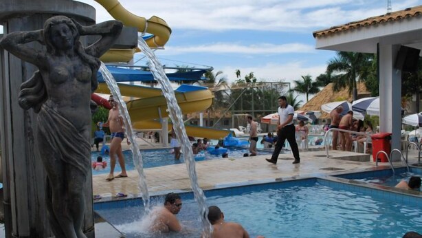A pool area with a water slide, a statue, and people swimming and relaxing. - Apartment diRoma Resort - Achei Férias (Caldas Novas)