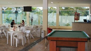 A pool table in a room with white chairs and tables, large windows, and a TV mounted on the wall. - Apartment diRoma Resort - Achei Férias (Caldas Novas)