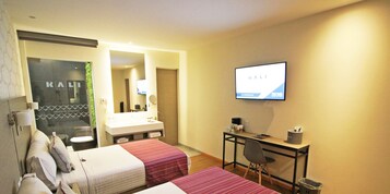 Habitación de hotel con dos camas, un escritorio, un televisor y un baño con espejo y lavamanos.