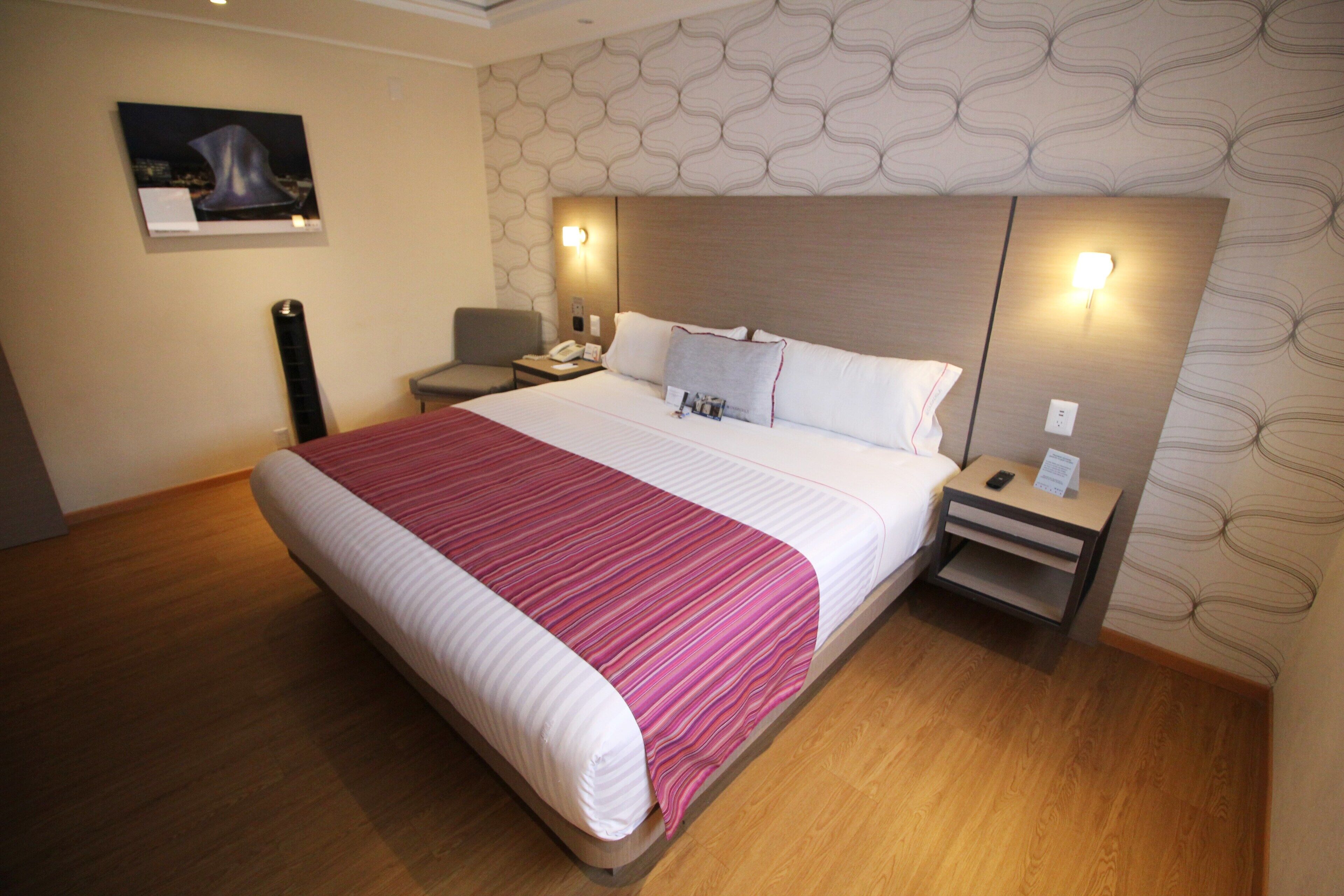 A hotel room with a large bed, a nightstand, a chair, and a wall-mounted light.