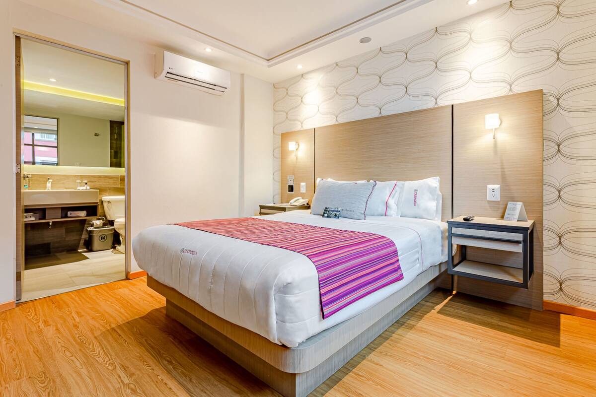 A modern hotel room with a large bed, a bedside table, a nightstand, a mirror, and a bathroom visible through an open door.