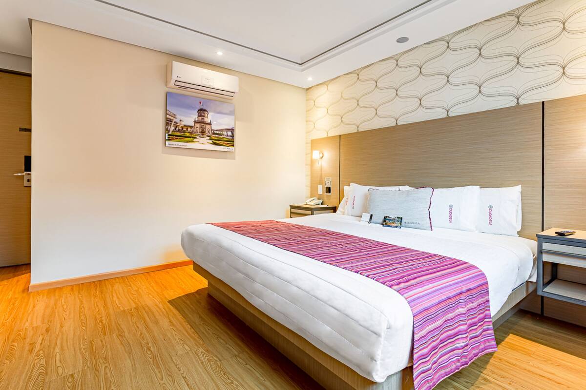 A modern hotel room with a large bed, wooden flooring, a decorative wall panel, an air conditioning unit, and a nightstand with a lamp.