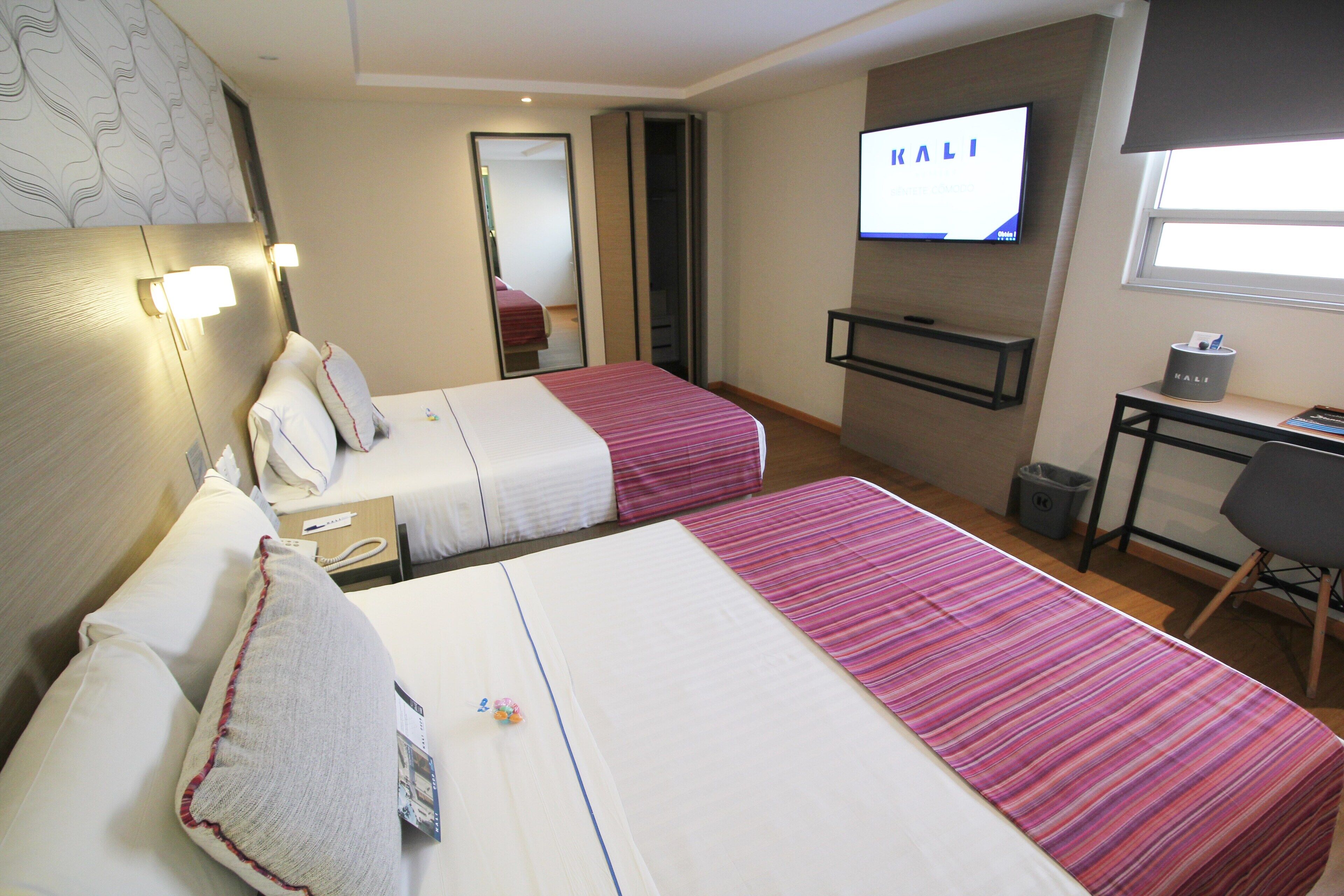 Ein Hotelzimmer mit zwei Betten, einem Fernseher, einem Schreibtisch und einem Spiegel.