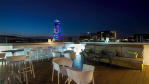 Un bar situé sur un toit, doté de mobilier moderne et offrant une vue sur les toits de la ville au crépuscule.