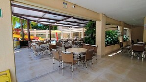 An outdoor cafe area with tables and chairs, a covered section, and a playground visible in the background. - Royal Residence - Achei Férias (Caldas Novas)