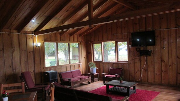 Una cabaña de madera con techo alto, un sofá, dos sillones, una mesa de centro, un televisor montado en la pared y ventanas amplias.