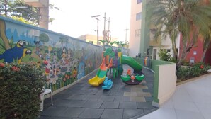 A children's play area with a slide and climbing structures, surrounded by a mural and palm trees. - Vitória das Thermas - Achei Férias (Caldas Novas)