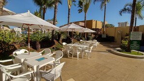 Un espace extérieur aménagé en terrasse, avec des tables et des chaises en plastique blanc, entouré de palmiers et d’une piscine.