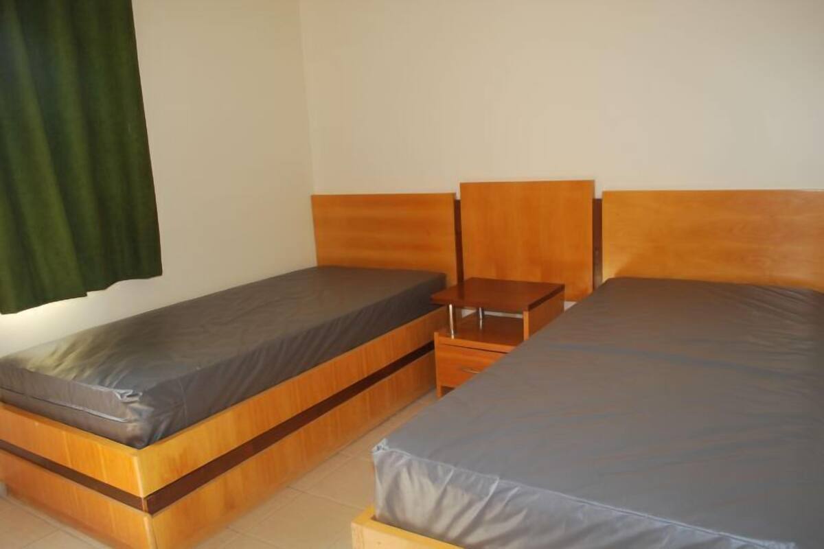 Una habitación individual con dos camas de madera, una mesita de noche de madera y una cortina verde.