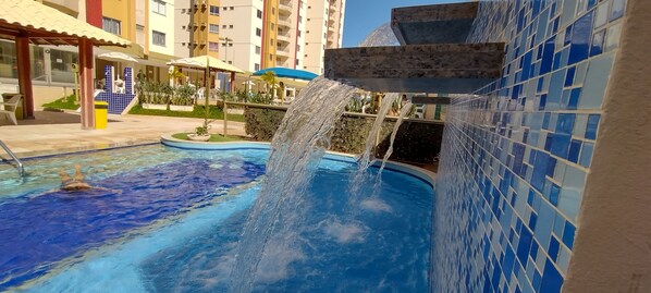 A swimming pool with a waterfall feature, surrounded by a tiled wall and a covered seating area.