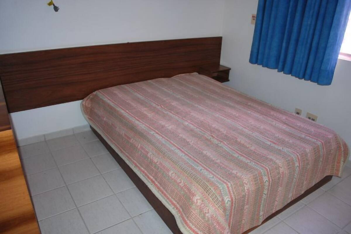 Un dormitorio con cabecera de madera, colcha estampada, mesita de noche y ventana con cortinas azules.