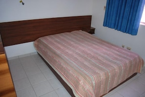 A bedroom with a wooden headboard, a patterned bedspread, a bedside table, and a window with blue curtains.