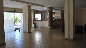 Eine großzügige Lobby mit Fliesenböden, hohen Säulen, einem Empfangsschalter und einer Glastür zum Außenbereich.