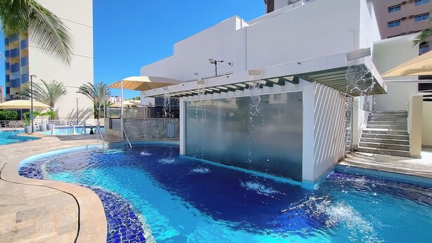 A swimming pool with a glass enclosure, surrounded by a modern building with a white facade and a covered area with umbrellas.