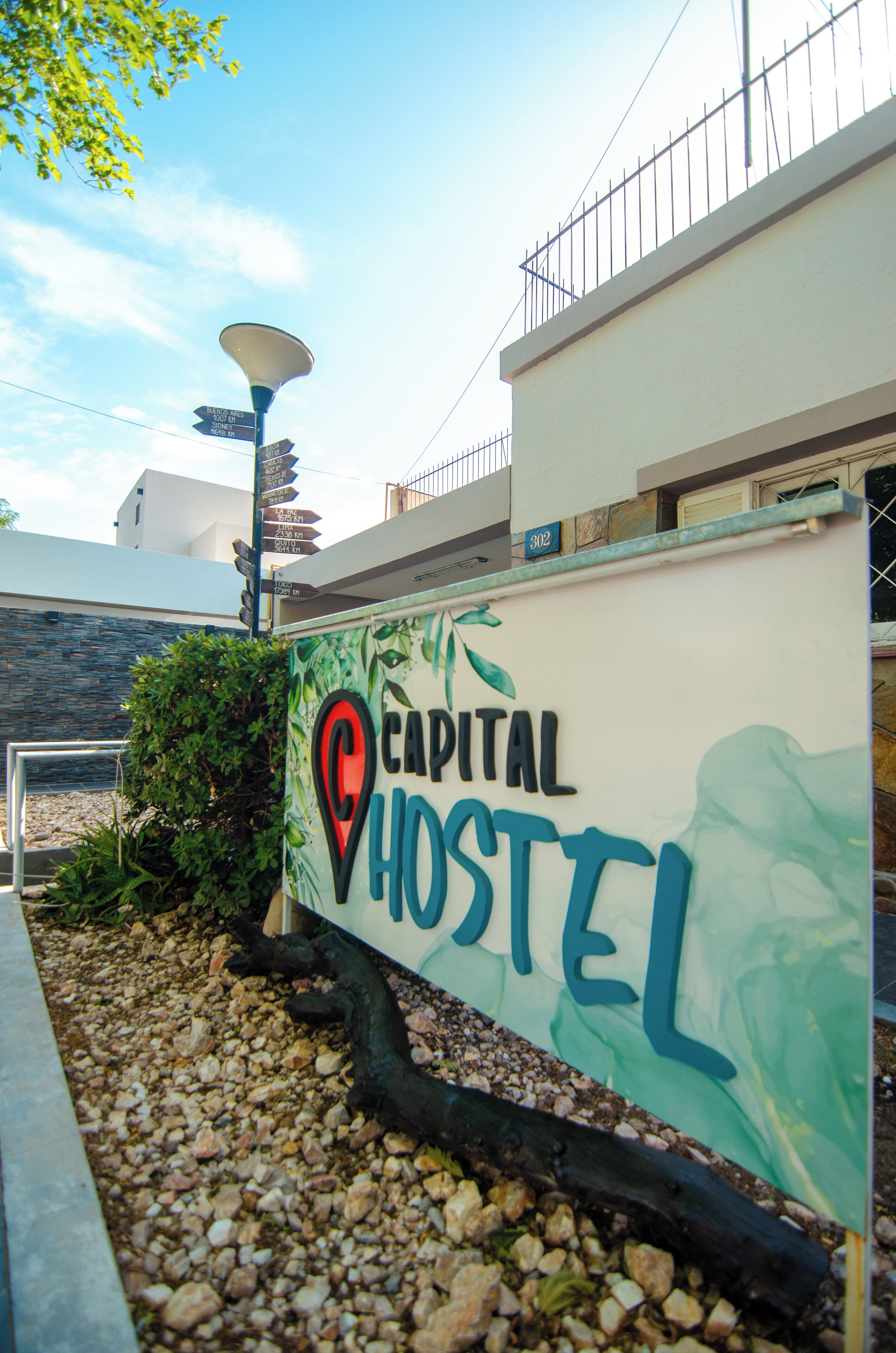 A hostel sign with a map pin logo, surrounded by pebbles and greenery.