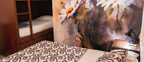 A bunk bed with a patterned bedspread and pillows, a desk with a lamp, and a wall with a floral painting.