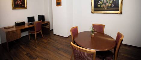 A room with a wooden desk, two chairs, a round table with a potted plant, and three framed artworks on the walls.