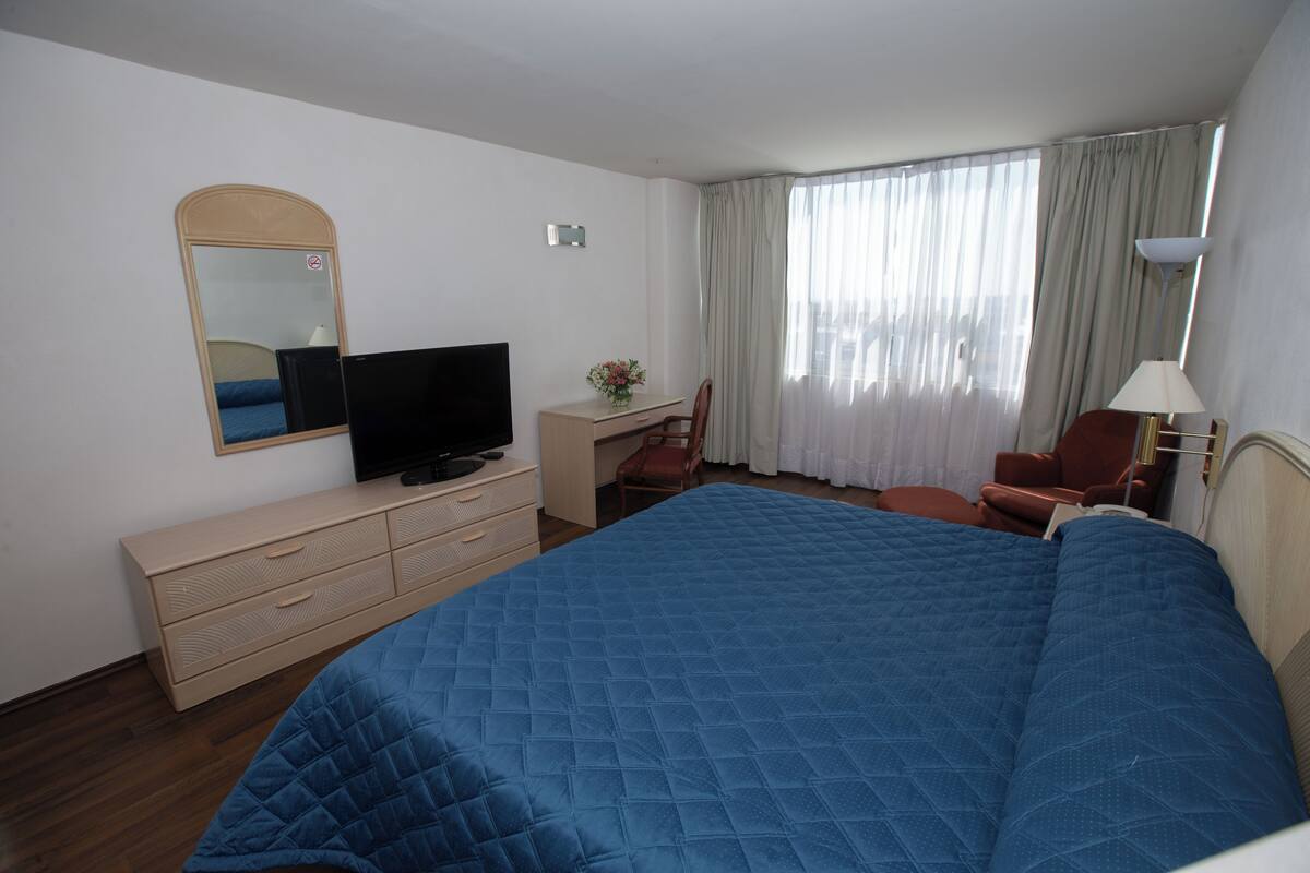 Ein Hotelzimmer mit einem großen Bett, einem Schreibtisch mit Stuhl, einem Fernseher und einem Fenster mit Vorhängen.