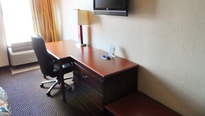 A hotel room with a wooden desk, a black office chair, a flat-screen TV mounted on the wall, a lamp, and a window with curtains. - Courtesy Inn (Oklahoma City)